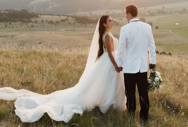 A bride and groom standing in a grassy field at sunset, holding hands and looking at each other.