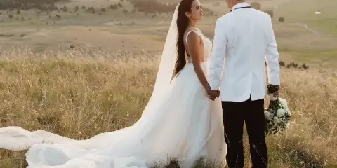 A bride and groom standing in a grassy field at sunset, holding hands and looking at each other.