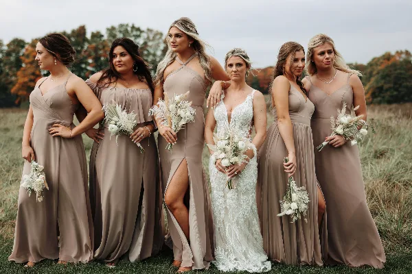 A bride in a white lace gown standing with five bridesmaids in beige dresses in a grassy field.