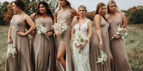 A bride in a white lace gown standing with five bridesmaids in beige dresses in a grassy field.