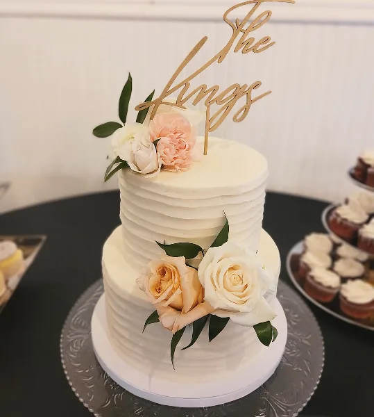 A two-tier white wedding cake with fresh floral decorations and a gold 'The Kings' topper.