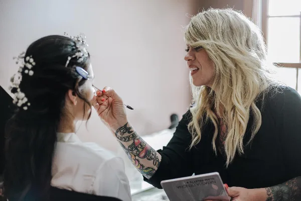 A makeup artist with blonde hair and tattoos applies makeup to a bride wearing a floral headpiece.