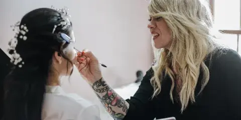 A makeup artist with blonde hair and tattoos applies makeup to a bride wearing a floral headpiece.