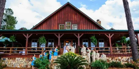 A rustic wooden lodge with a stone foundation and a wedding party posing on the front steps.