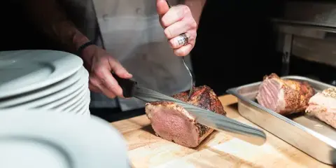 Chef carving a roasted meat on a wooden cutting board with stacked white plates nearby
