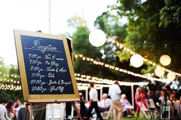 Outdoor wedding reception with a chalkboard schedule and string lights.