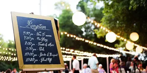 Outdoor wedding reception with a chalkboard schedule and string lights.