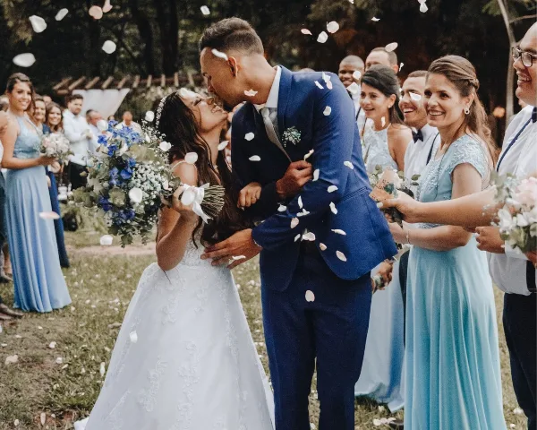 A bride and groom share a kiss during an outdoor wedding ceremony while guests throw flower petals.