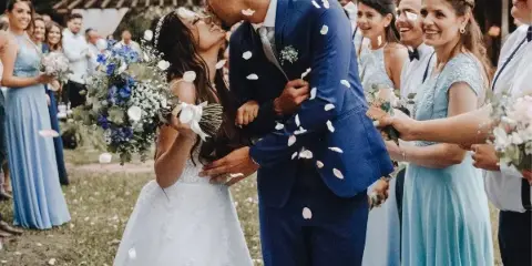 A bride and groom share a kiss during an outdoor wedding ceremony while guests throw flower petals.