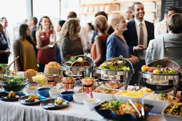 A catering buffet table filled with various food dishes and chafing dishes, with people standing in the background.