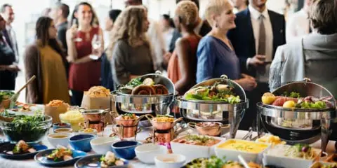 A catering buffet table filled with various food dishes and chafing dishes, with people standing in the background.