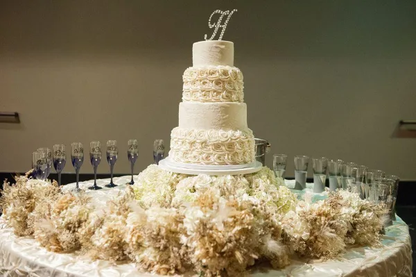 A four-tiered white wedding cake with ruffled floral details and a monogram topper displayed on a table with floral arrangements.