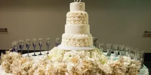 A four-tiered white wedding cake with ruffled floral details and a monogram topper displayed on a table with floral arrangements.