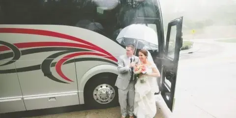 A bride and groom standing in front of a white shuttle bus with red and black graphics.