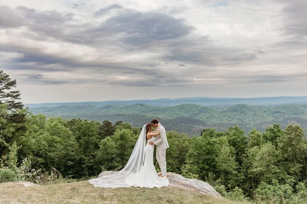 A bride and groom standing on a rocky ledge with a backdrop of rolling green hills and mountains.