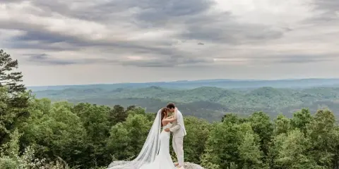 A bride and groom standing on a rocky ledge with a backdrop of rolling green hills and mountains.