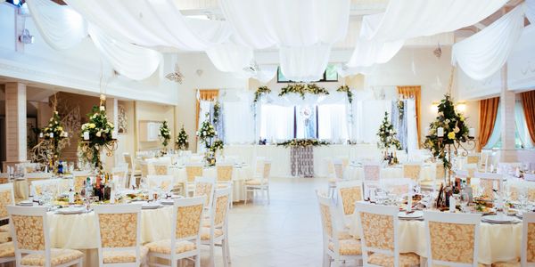 A spacious, elegantly decorated wedding reception hall with white tablecloths, floral centerpieces, and draped ceiling fabric.