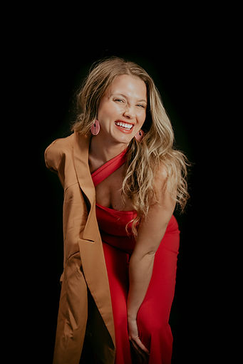 Portrait of a smiling woman with long blonde hair wearing a red dress and tan blazer against a black background.