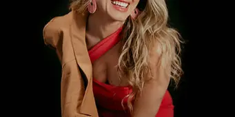 Portrait of a smiling woman with long blonde hair wearing a red dress and tan blazer against a black background.