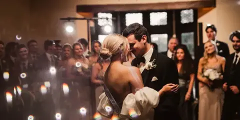 A bride and groom sharing a first dance at a wedding reception, surrounded by guests and wedding party members.
