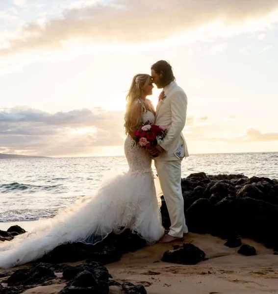 A bride and groom standing close together on a rocky beach at sunset, with the bride holding a bouquet of red and pink roses.