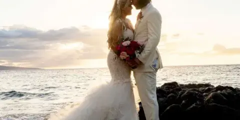 A bride and groom standing close together on a rocky beach at sunset, with the bride holding a bouquet of red and pink roses.