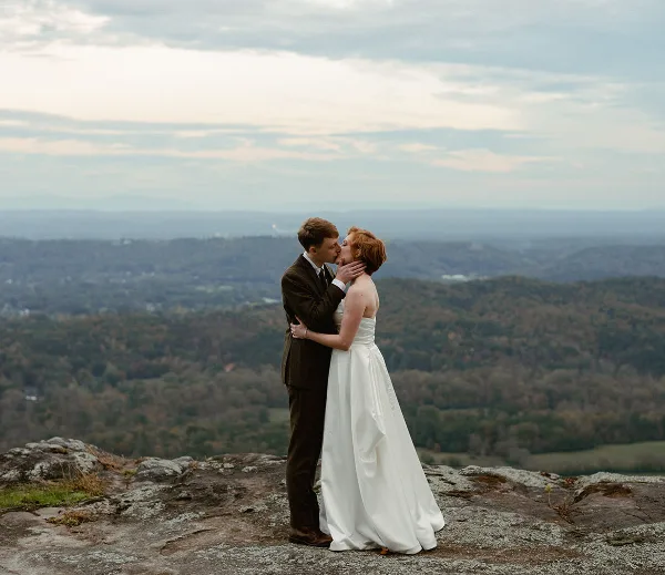 A bride and groom kissing on a rocky overlook with a vast landscape in the background.
