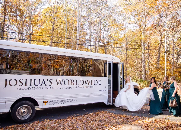 A white motor coach branded with 'Joshua's Worldwide' and 'Ground Transportation' text, with a bride and bridesmaids boarding the vehicle in an autumn setting.