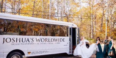 A white motor coach branded with 'Joshua's Worldwide' and 'Ground Transportation' text, with a bride and bridesmaids boarding the vehicle in an autumn setting.