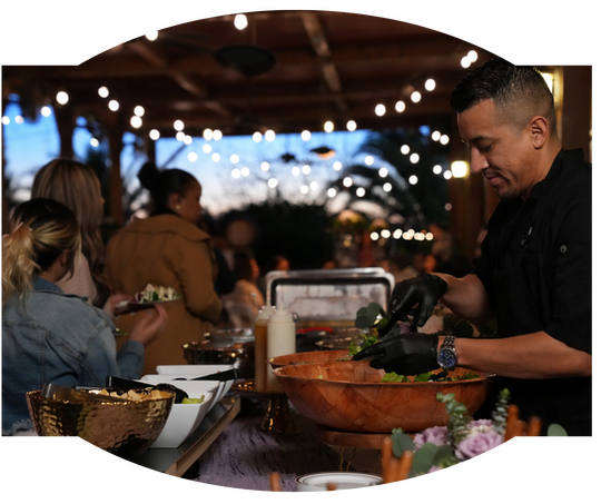 A catering staff member in a black shirt and gloves prepares a salad at a buffet table during an event.
