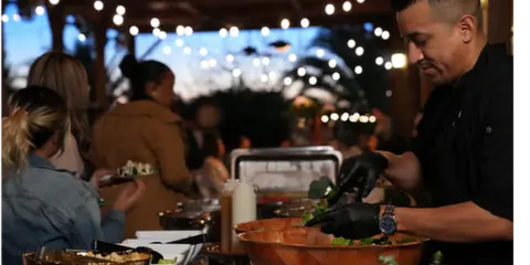A catering staff member in a black shirt and gloves prepares a salad at a buffet table during an event.