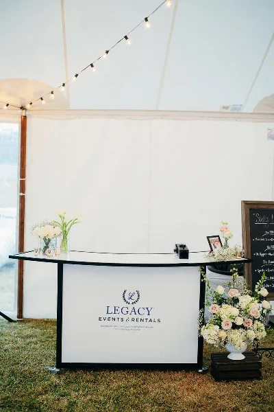 A white event table with a Legacy Events & Rentals branded skirt, decorated with flowers and string lights inside a white tent.