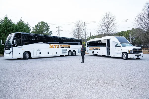 A man in a suit standing in a parking lot between a large white charter bus and a white shuttle van, both featuring MTI Limo and Shuttle Services branding.
