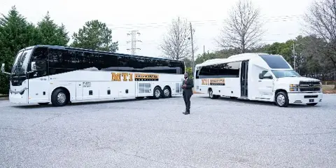 A man in a suit standing in a parking lot between a large white charter bus and a white shuttle van, both featuring MTI Limo and Shuttle Services branding.