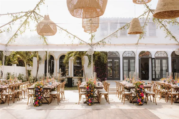 Outdoor wedding reception venue with long tables, vibrant floral centerpieces, and woven pendant lights under a white tent.