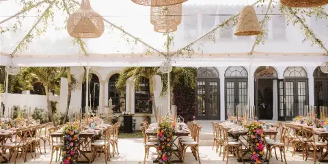 Outdoor wedding reception venue with long tables, vibrant floral centerpieces, and woven pendant lights under a white tent.
