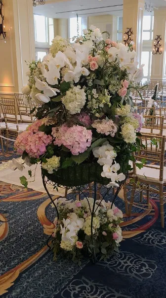 A tall, lush floral arrangement featuring white orchids, pink and white hydrangeas, and greenery, displayed on a stand in a formal event venue.
