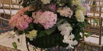 A tall, lush floral arrangement featuring white orchids, pink and white hydrangeas, and greenery, displayed on a stand in a formal event venue.