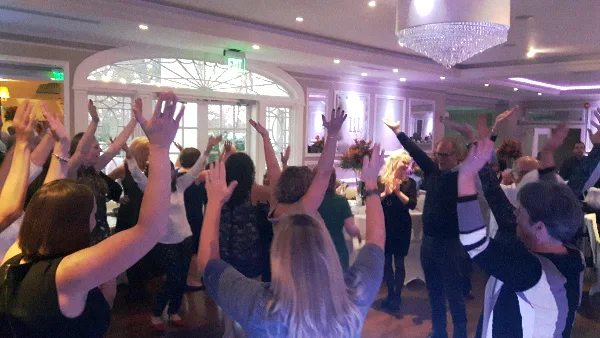 Guests dancing at a wedding reception with raised hands under colored event lighting and a chandelier.
