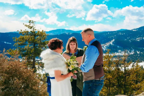 A wedding officiant performing a ceremony for a bride and groom on a mountain overlook with a scenic background.