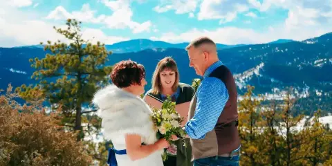 A wedding officiant performing a ceremony for a bride and groom on a mountain overlook with a scenic background.