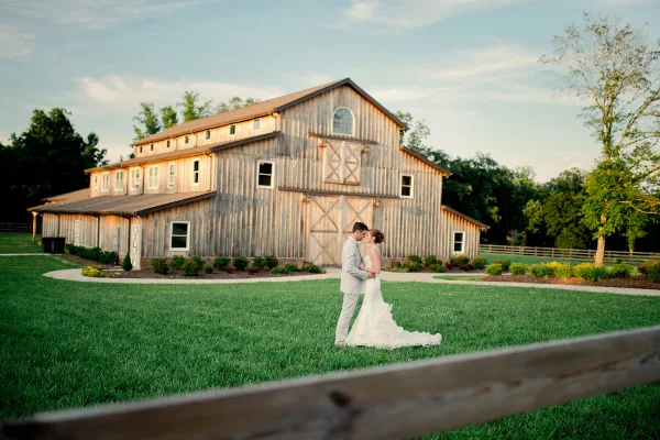 A bride and groom standing on a green lawn in front of a large, rustic wooden wedding barn.