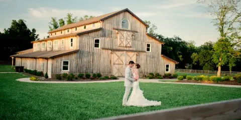 A bride and groom standing on a green lawn in front of a large, rustic wooden wedding barn.