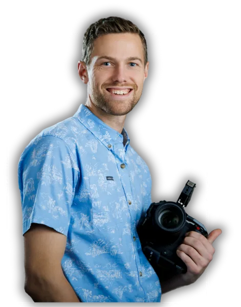 Portrait of a smiling man in a blue patterned shirt holding a professional video camera against a black background.
