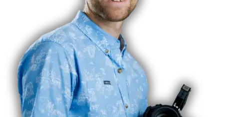 Portrait of a smiling man in a blue patterned shirt holding a professional video camera against a black background.