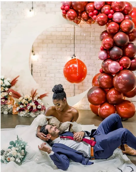 A bride and groom posing in front of a large red balloon arch and floral arrangements.