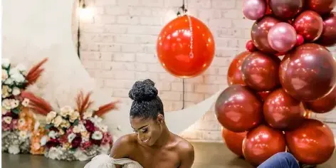 A bride and groom posing in front of a large red balloon arch and floral arrangements.