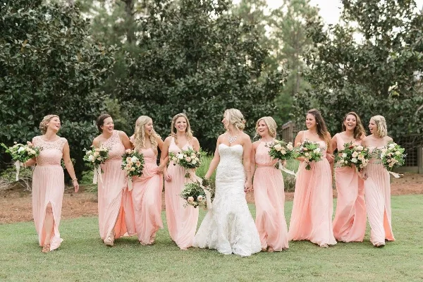 A bride and bridesmaids in peach dresses holding floral bouquets outdoors.