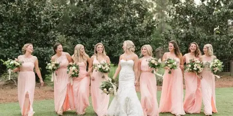 A bride and bridesmaids in peach dresses holding floral bouquets outdoors.