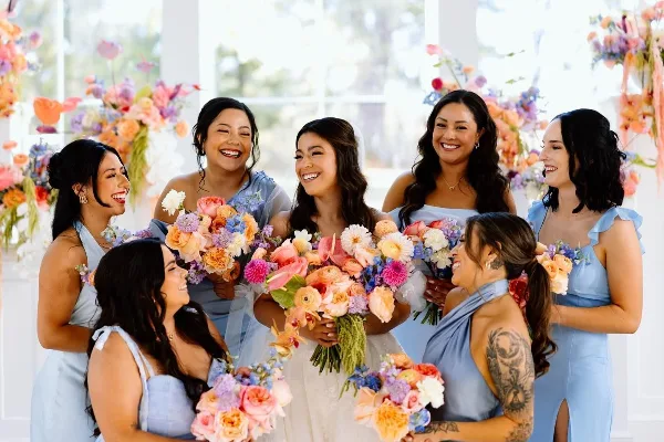 Bride and bridesmaids in light blue dresses smiling with colorful bouquets at a wedding.